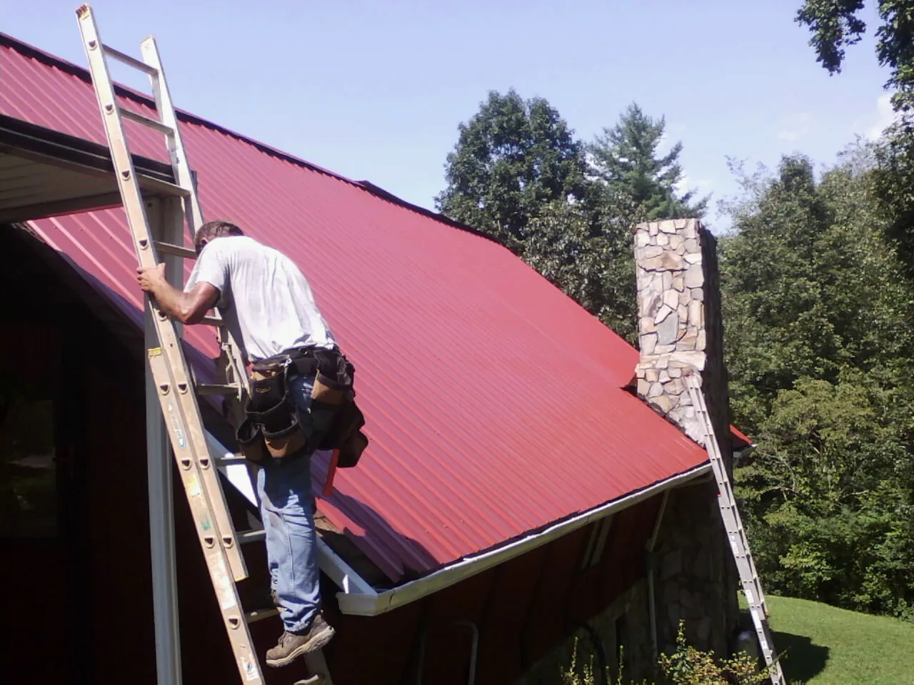 Roofer on a ladder beside a red metal roof and stone chimney — Soffit & Fascia Repair by Roofing Brothers in Elburn